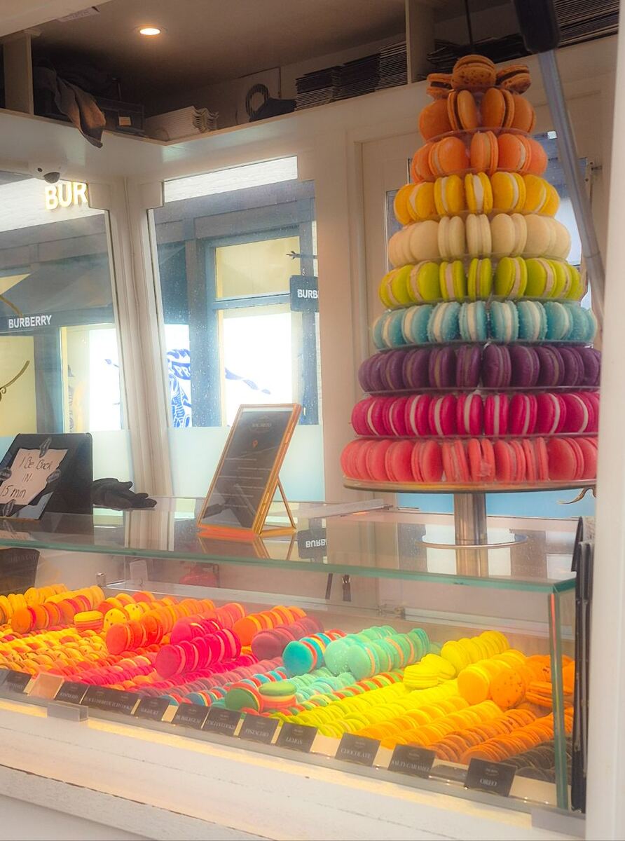 Een vitrine vol kleurrijke macarons in een winkel, met een grote toren van macarons in regenboogkleuren ernaast. Op de achtergrond is door het raam een gebouw met het logo 'Burberry' te zien.