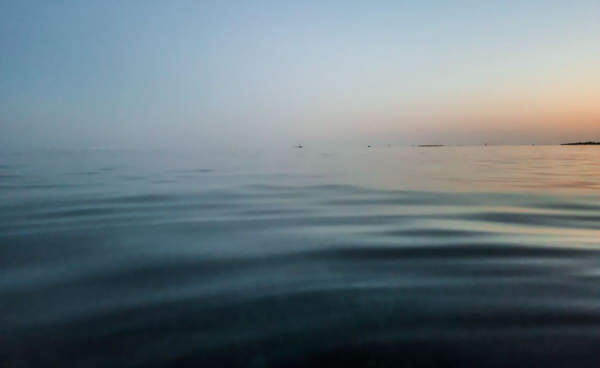 De foto toont een rustig wateroppervlak met lichte golvingen, gezien vanaf een laag perspectief. De horizon is zichtbaar met een zachte overgang van blauw naar een oranje gloed aan de rechterkant, wat duidt op zonsopkomst of zonsondergang. In de verte zijn enkele kleine objecten op het water te zien, waaronder een boei en een vaartuig.
