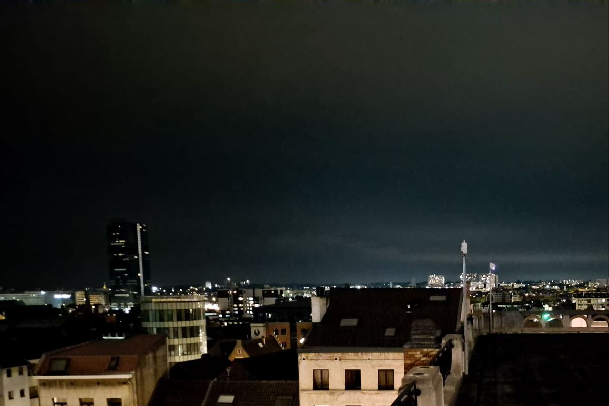 Een nachtelijk stadsgezicht met op de voorgrond daken van gebouwen en op de achtergrond de verlichte skyline van een stad met een opvallende hoge toren.