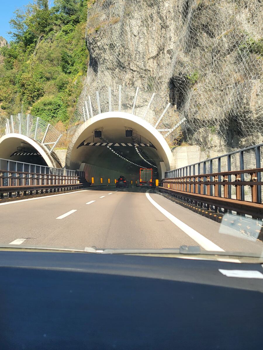 Een snelweg die leidt naar twee tunnelmonden, omgeven door steile, rotsachtige bergwanden. De bergwanden zijn deels begroeid met groen en voorzien van veiligheidsnetten. In de rechtertunnel rijden een auto en een vrachtwagen voor ons uit. De foto is genomen vanuit een voertuig en toont de voorruit en het dashboard aan de onderkant.