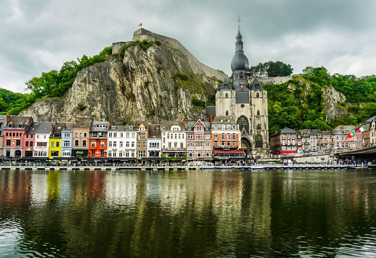 Een breed overzicht van de stad Dinant, gezien vanaf het water. Op de voorgrond is de Maas te zien met de weerspiegeling van de gebouwen. Langs de oever staan kleurrijke huizen en restaurants met terrassen. Rechts van het midden staat de Collegiale kerk van Onze-Lieve-Vrouw met zijn opvallende peervormige toren. Achter de gebouwen rijst een steile rotswand op, met daarop de citadel van Dinant.