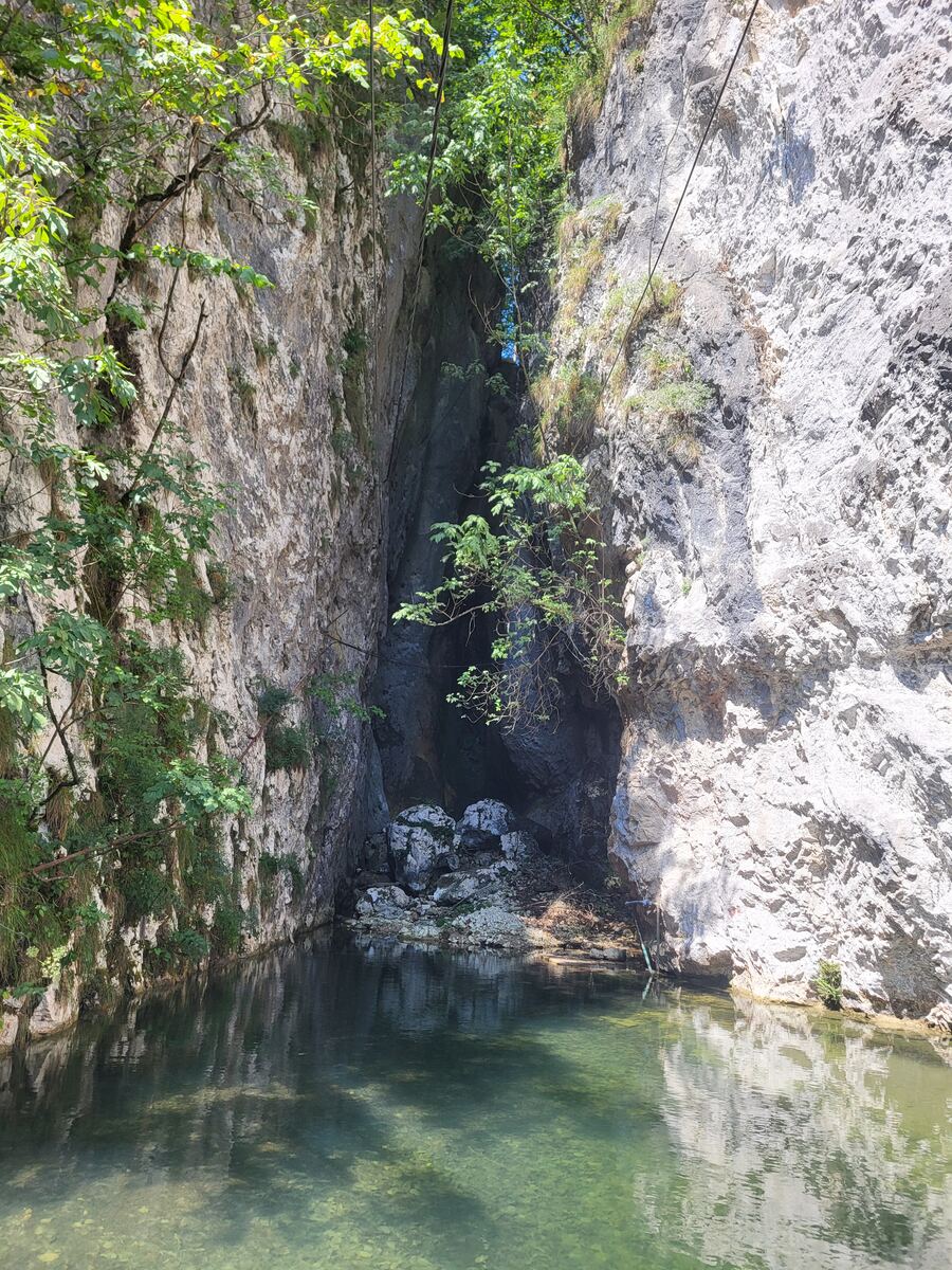 Een diepe kloof met steile rotswanden en helder groen water op de voorgrond. Bovenin de kloof groeien bomen en struiken, en er zijn enkele kabels zichtbaar die over de kloof gespannen lijken te zijn. De zon schijnt op de rotswanden en het water.