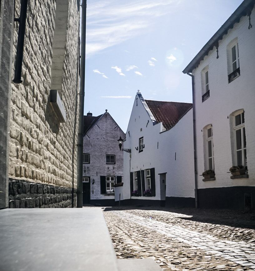 Een smal straatje met kinderkopjes, omringd door witgepleisterde huizen met donkere luiken en rode daken. De zon schijnt fel en werpt lange schaduwen op de straat. De lucht is blauw met enkele witte wolkjes.