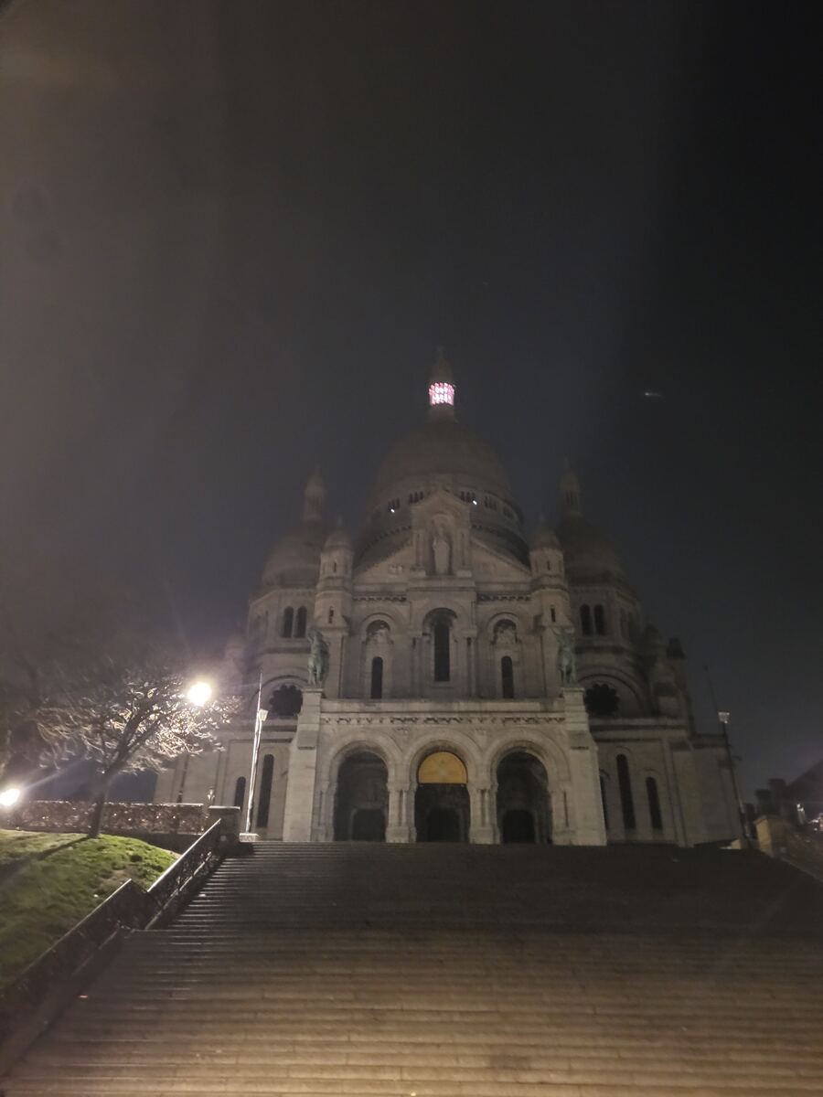 Een groots gebouw met koepels en torens, vermoedelijk een basiliek, staat verlicht tegen een donkere nachtelijke of zeer vroege ochtendhemel. Een lange, brede stenen trap leidt omhoog naar de ingang van het gebouw. Aan de linkerzijde is een kale boom zichtbaar, samen met een felle straatlantaarn die licht werpt op de omgeving. De lucht is donker en lijkt licht nevelig. Er zijn geen mensen zichtbaar op de trappen of rond het gebouw.