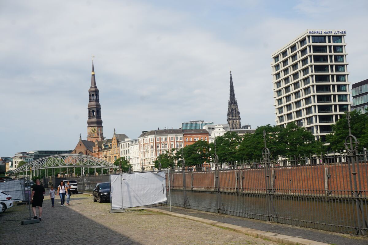 Een stadsgezicht met op de voorgrond een kade langs water, afgeschermd door een hek. Op de achtergrond zijn diverse gebouwen te zien, waaronder twee kerktorens en een modern wit gebouw met de tekst 'MOHRLE HAPP LUTHER'. Er lopen enkele mensen op de kade en er staan auto's geparkeerd.
