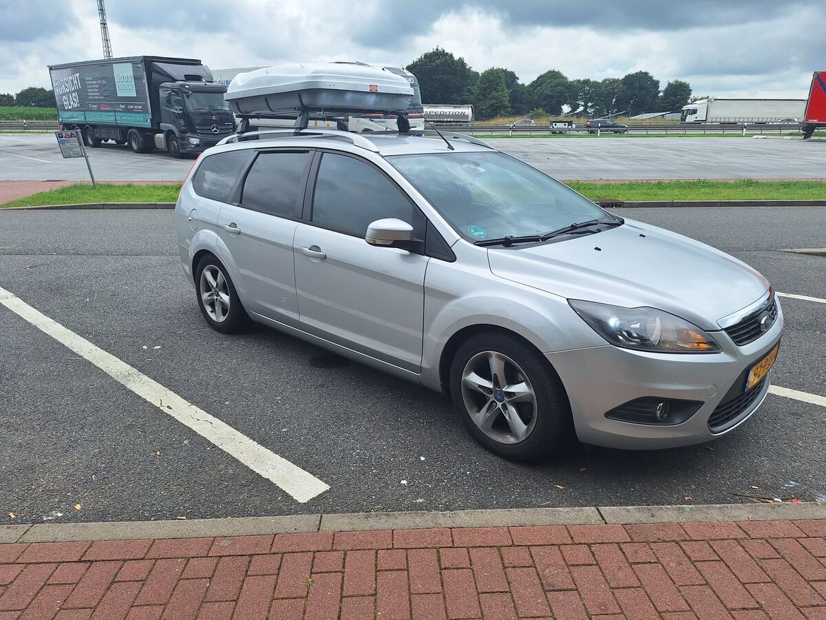 Een zilverkleurige Ford Focus stationwagen met een dakkoffer staat geparkeerd op een parkeerplaats langs de snelweg. Op de achtergrond zijn vrachtwagens en bomen te zien onder een bewolkte lucht. De auto staat op een asfaltparkeerplaats met witte belijning en een strook rode klinkers op de voorgrond.