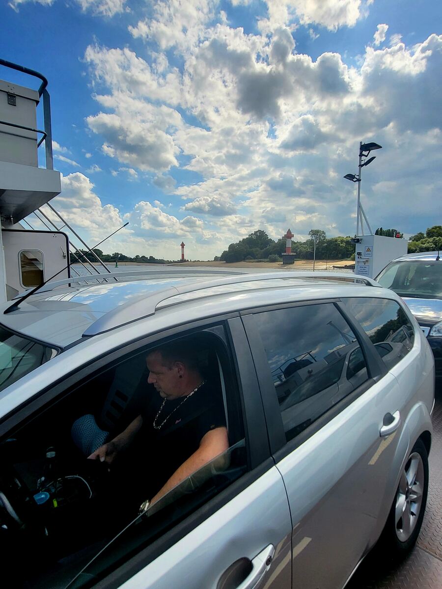 Een man zit in een zilverkleurige Ford Focus stationwagen op een veerboot. Op de achtergrond zijn twee rood-wit gestreepte vuurtorens te zien, met een zandstrand en bomen langs de waterkant onder een blauwe lucht met witte wolken.