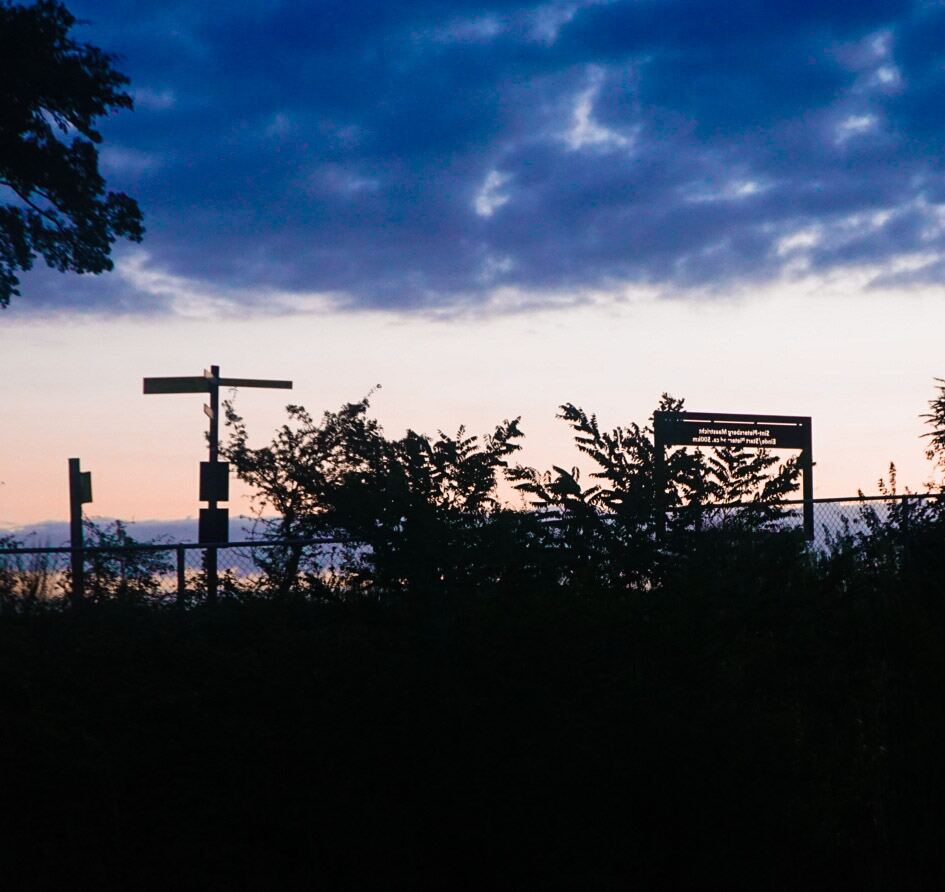 Een silhouet van bomen en een hek tegen een avondlucht met donkerblauwe wolken en een oranje gloed aan de horizon. Er zijn twee palen zichtbaar, waarvan één met een dwarsbalk en de andere met een rechthoekig bord met onleesbare tekst.