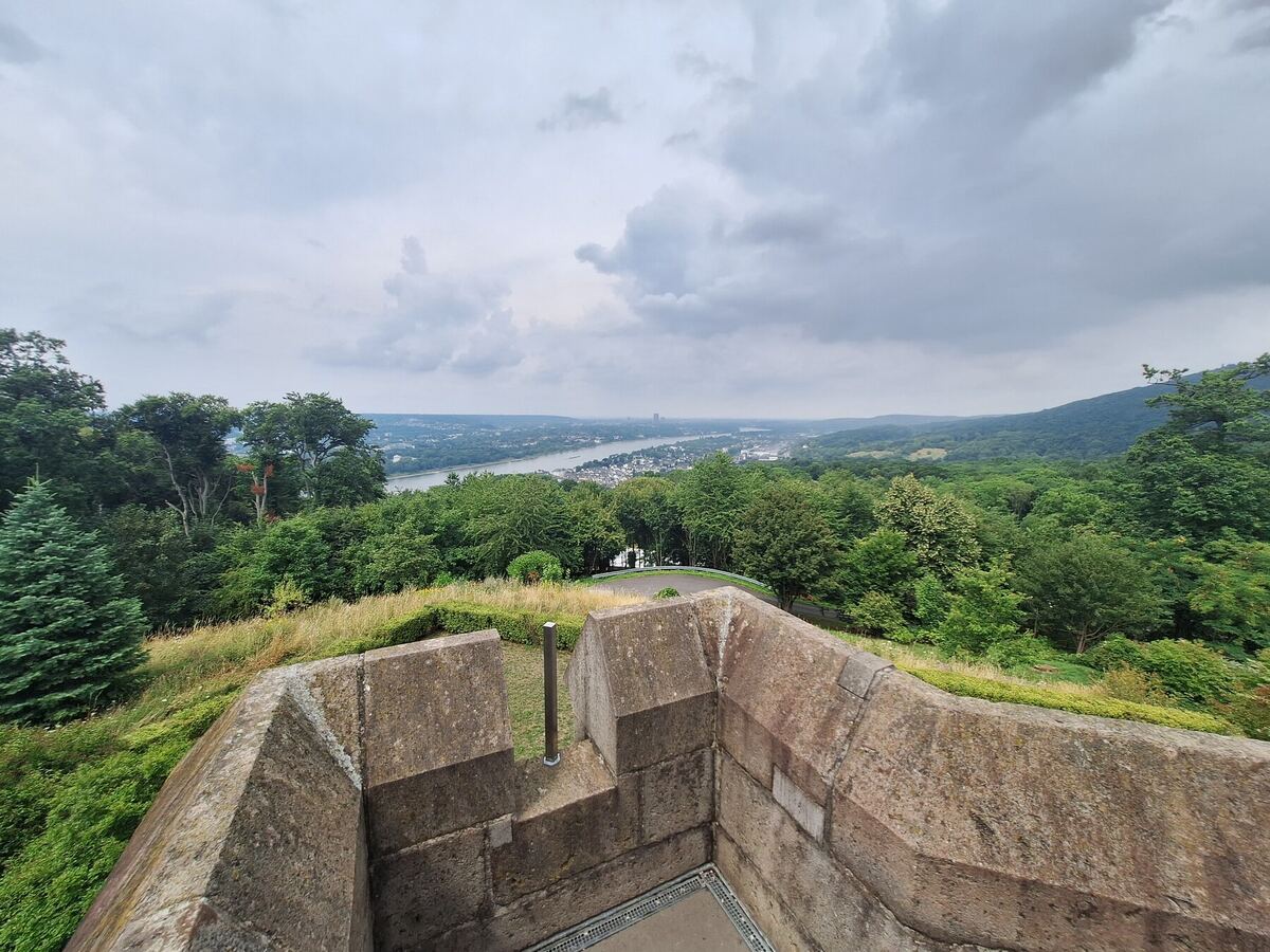 Een weids uitzicht over een groene vallei met een rivier, gezien vanaf een stenen uitkijkpunt. De lucht is bewolkt en de horizon is gevuld met beboste heuvels en een stad langs de rivier.