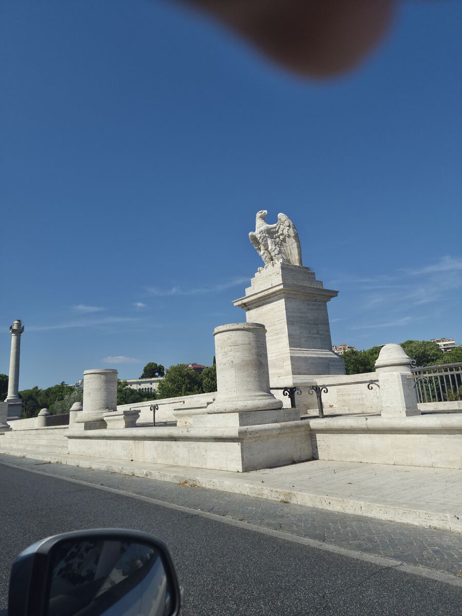 Een grote witte stenen adelaar op een hoge sokkel, onderdeel van een monumentale brug met klassieke zuilen en een balustrade. Op de voorgrond is een weg en de zijspiegel van een auto zichtbaar onder een helderblauwe lucht met enkele wolkjes. Op de achtergrond zijn groene bomen en gebouwen te zien.