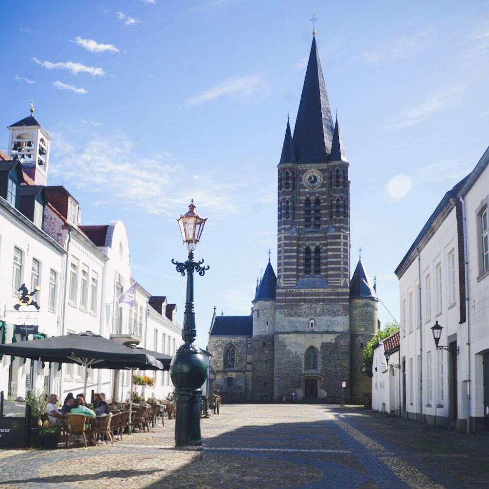 Een zonnig kerkplein met een grote, donkere kerktoren in het midden, omringd door witte gebouwen met rode daken. Links is een terras met zwarte parasols en mensen die buiten zitten. Op de voorgrond staat een sierlijke lantaarnpaal.