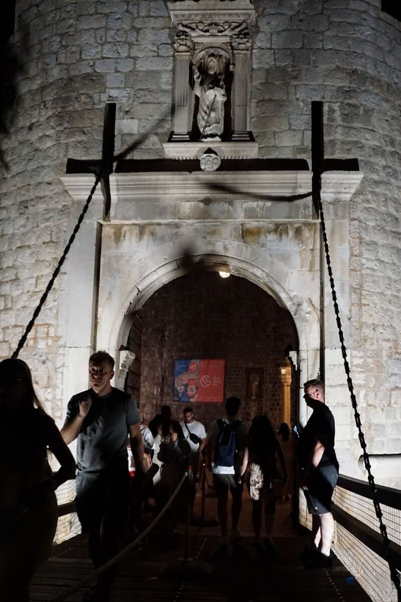Een foto genomen bij nacht van de Pilepoort in Dubrovnik, met een stenen boog en een standbeeld van Sint Blasius erboven. Mensen lopen over de houten brug die naar de poort leidt. De scène is verlicht door kunstlicht, waardoor schaduwen ontstaan.