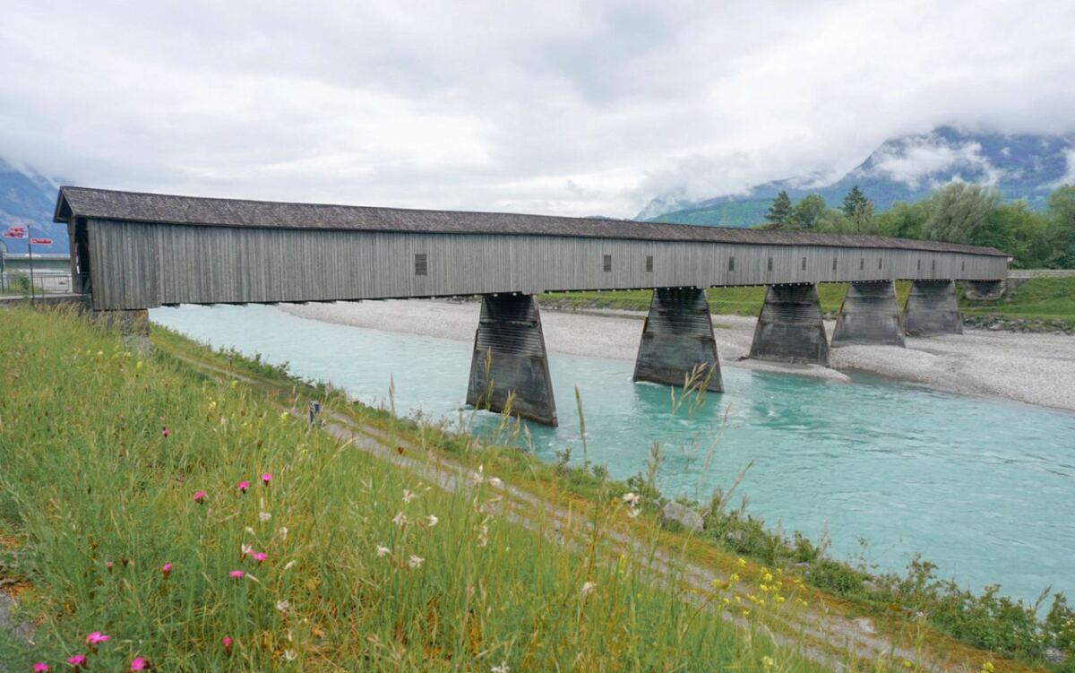 Een lange, houten overdekte brug met een pannendak, ondersteund door betonnen pilaren, overspant een turquoise rivier. Op de achtergrond zijn bergen en bomen te zien onder een bewolkte lucht. Op de voorgrond is een groene oever met wilde bloemen en een smal pad zichtbaar.