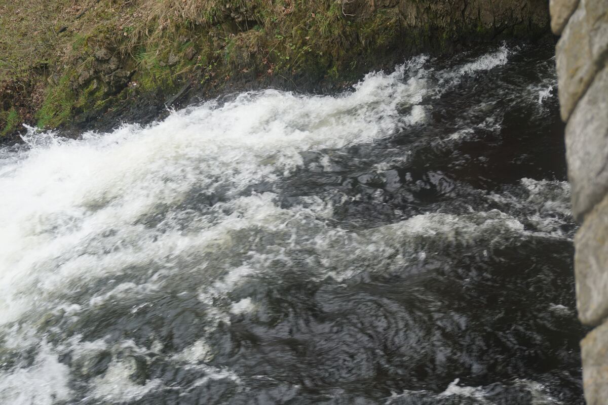 Een close-up van snelstromend water onder een brug, met schuimende witte golven en donkere waterpartijen. Aan de linkerkant is een begroeide oever zichtbaar en rechts een deel van een stenen brugleuning.