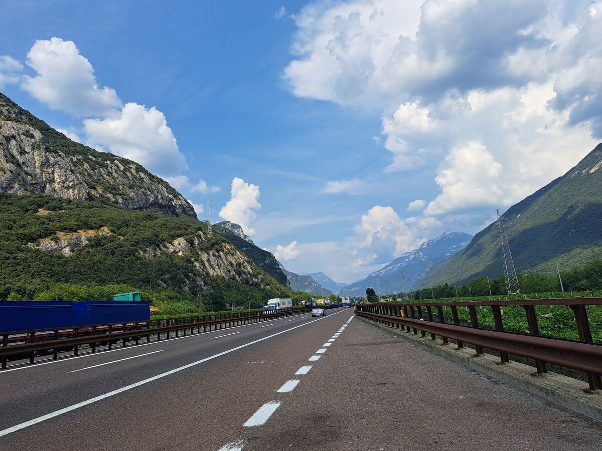 Een snelweg, de Autostrada del Brennero, snijdt door een bergachtige vallei onder een helderblauwe lucht met verspreide witte wolken. Links en rechts van de weg rijzen steile, deels rotsachtige en deels dichtbegroeide bergwanden op. In de verte zijn nog meer bergen zichtbaar. Op de snelweg rijden enkele personenauto's en vrachtwagens. Aan de rechterkant van de weg zijn vangrails en een hekwerk te zien, met daarachter groene vegetatie en enkele elektriciteitsmasten.