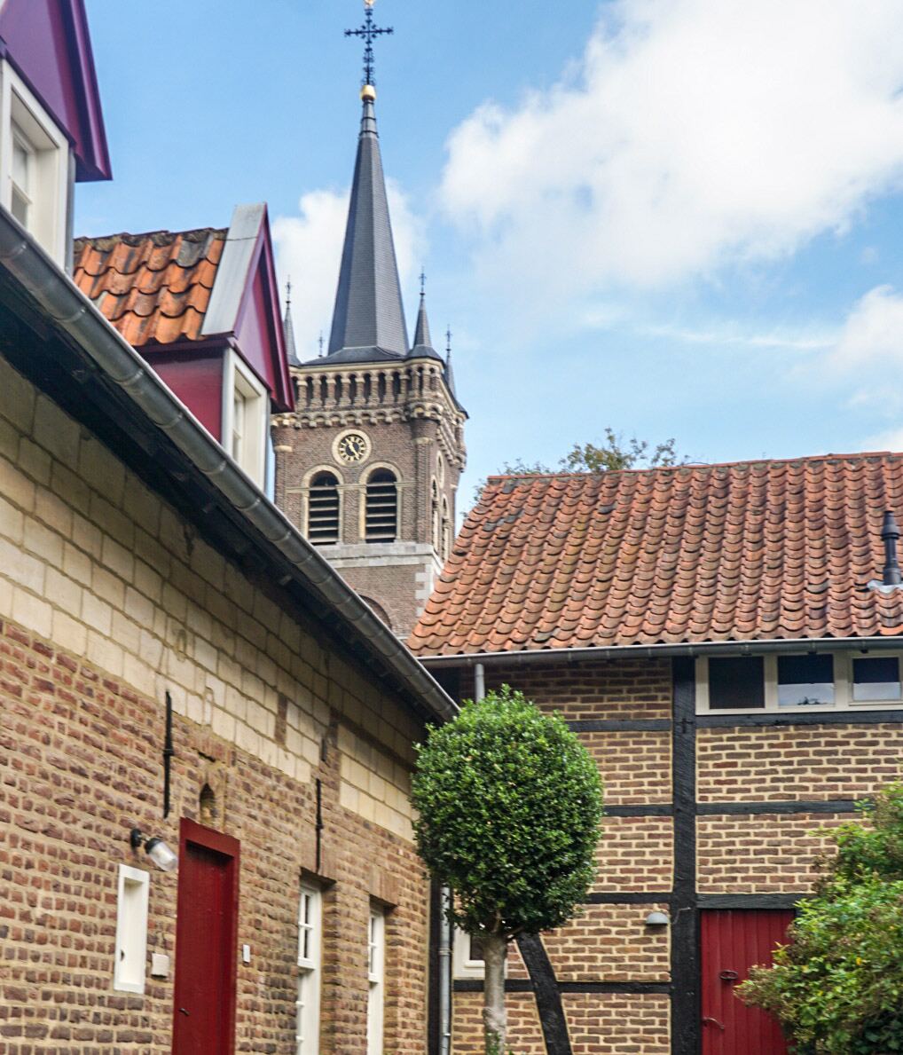Een foto van een kerktoren die boven de daken van oude bakstenen huizen uitsteekt, met een blauwe lucht en wolken op de achtergrond. De huizen hebben rode dakpannen en een van de huizen heeft vakwerk. Een klein boompje staat in het midden van de foto.