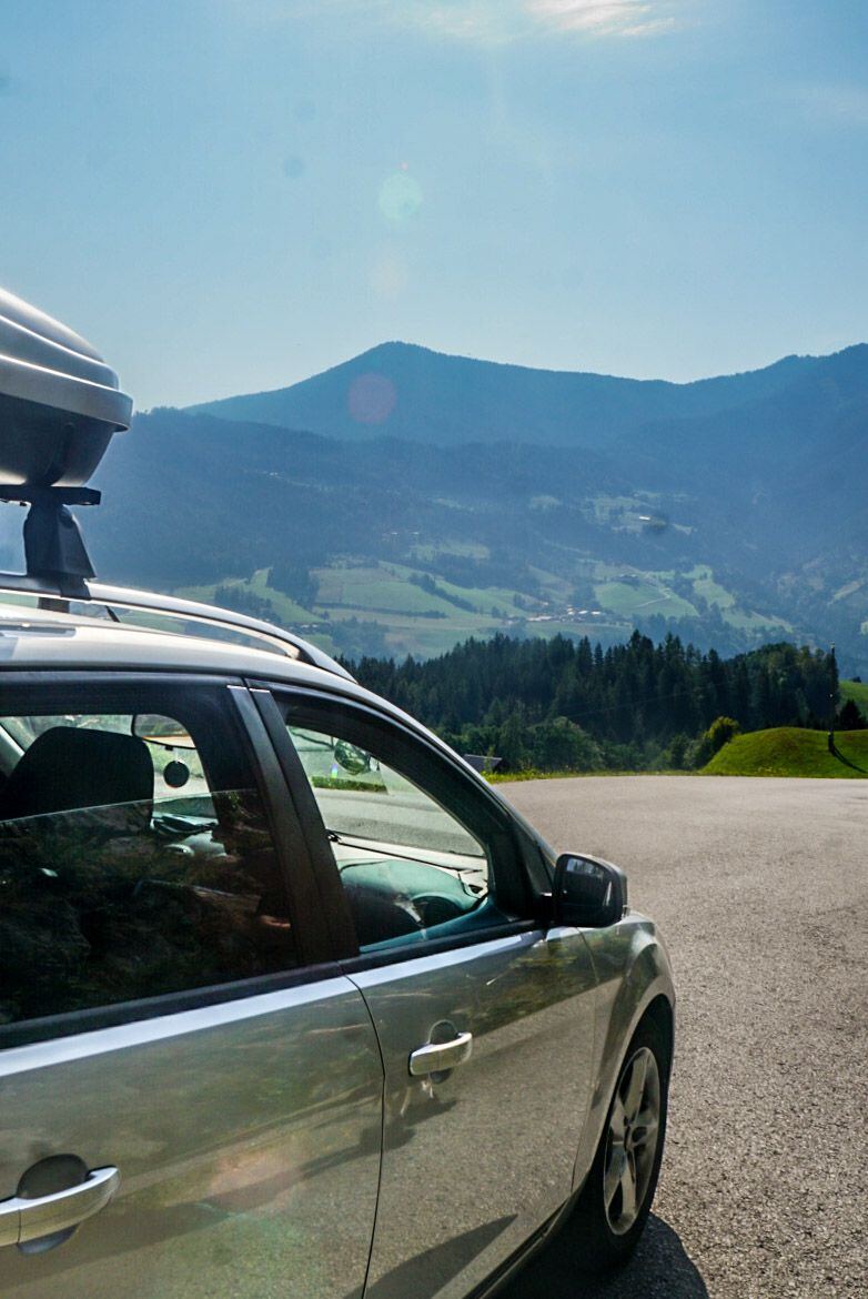 Een zilverkleurige Ford Focus stationwagen met een dakkoffer staat geparkeerd op een asfaltvlakte, met op de achtergrond een bergachtig landschap onder een helderblauwe lucht. De zon schijnt fel en veroorzaakt lensflares. De bergen zijn bedekt met bossen en groene weiden, en in de verte zijn enkele huizen zichtbaar.