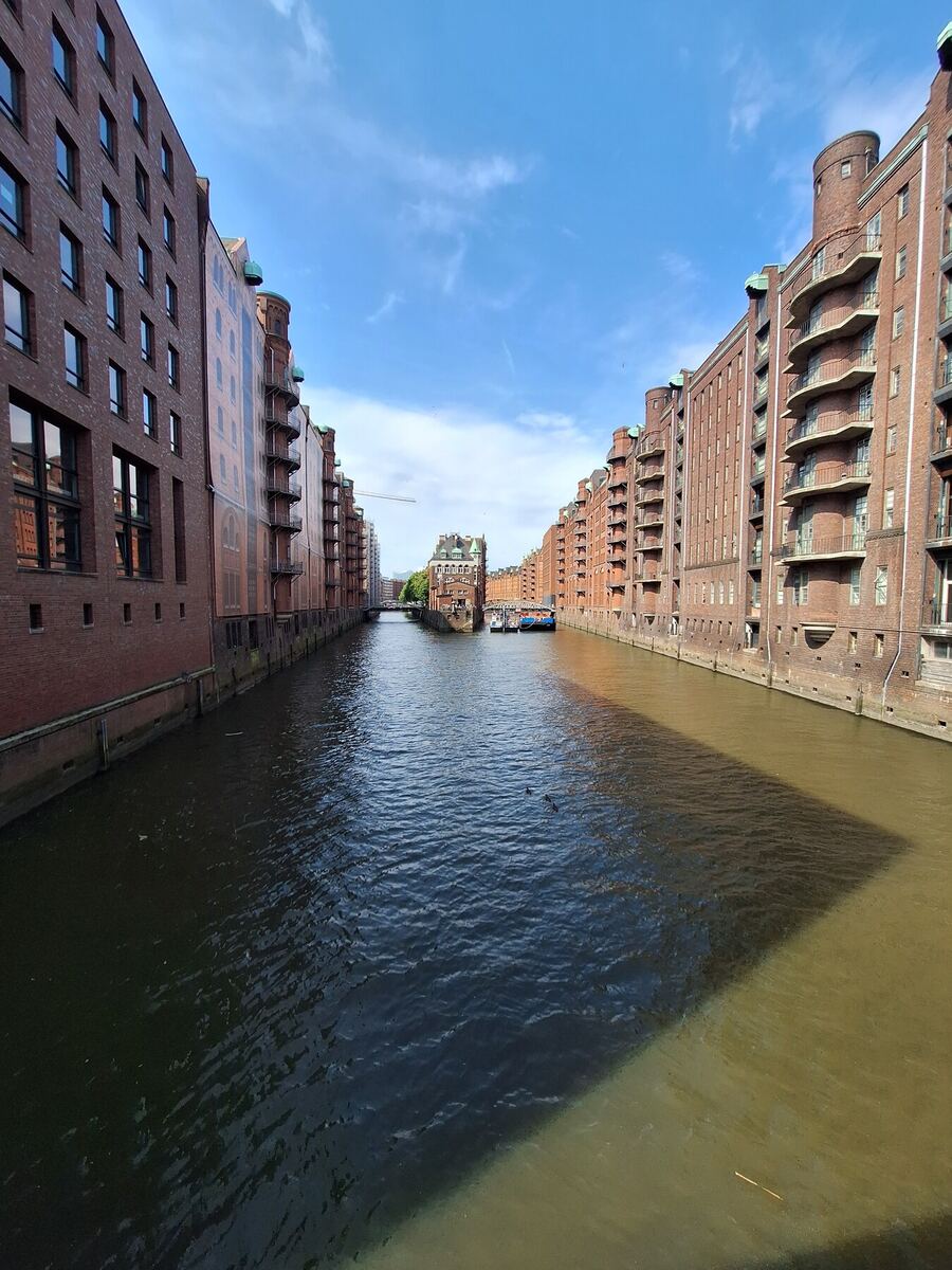 Een brede gracht met aan weerszijden hoge, roodbruine bakstenen pakhuizen onder een helderblauwe lucht. In de verte is een brug zichtbaar en een gebouw met een groen dak. Het water is deels in de schaduw en deels verlicht door de zon.