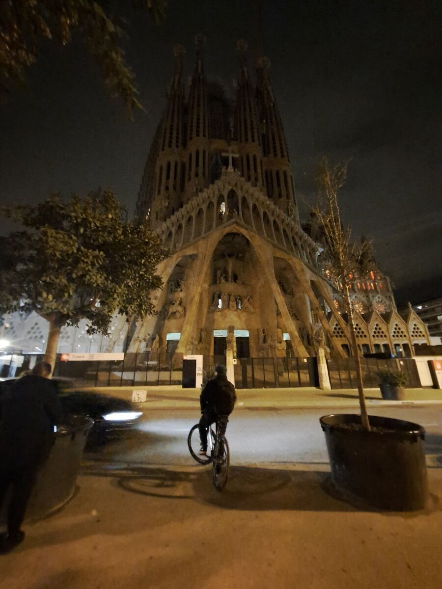 Een avondopname van de Sagrada Família in Barcelona, met een fietser op de voorgrond en een persoon aan de linkerkant. De basiliek is deels verlicht en de lucht is donker.