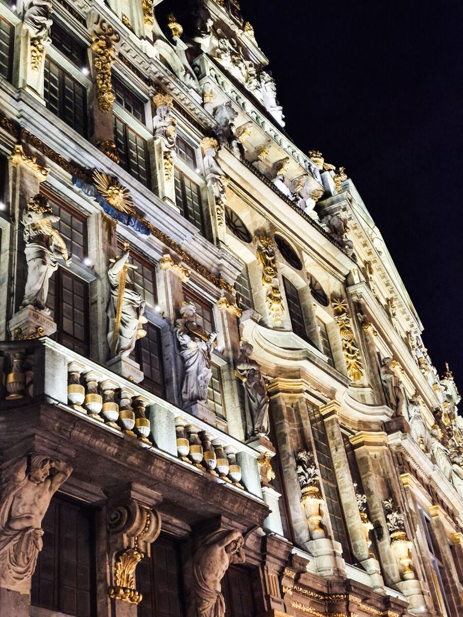 Een close-up van een rijk versierde, klassieke gevel van een gebouw, verlicht in het donker. De gevel heeft meerdere verdiepingen met ramen, pilasters, beelden en gouden ornamenten. De foto is genomen vanuit een laag standpunt, schuin omhoog kijkend naar de architectuur.