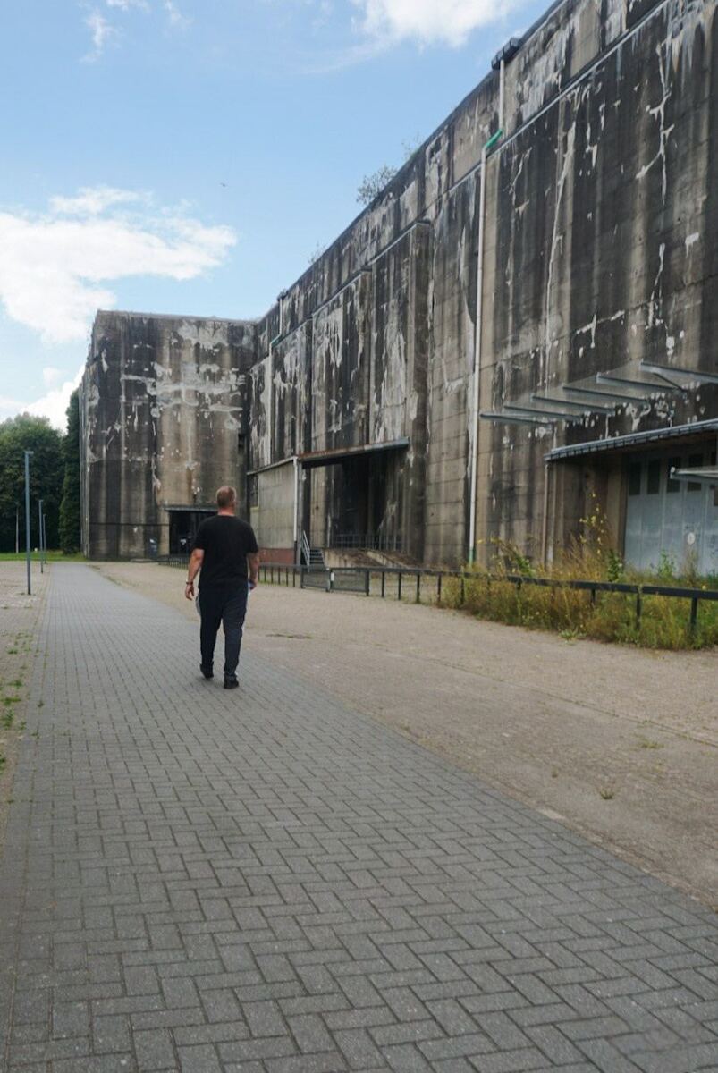 Een man loopt over een geplaveid pad richting een enorme betonnen bunker met een donkere, verweerde uitstraling. De bunker heeft hoge muren met verticale strepen en er zijn enkele kleine gebouwen of aanbouwen aan de zijkant te zien. De lucht is deels bewolkt en er staan bomen aan de linkerkant van het pad.