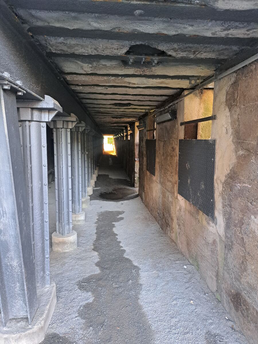 Een lange, rechte gang of onderdoorgang, gezien vanuit een laag perspectief. Aan de linkerkant staat een rij donkergrijze, robuuste metalen pilaren met klinknagels, die een zwaar, betonnen plafond lijken te dragen. Het plafond is ruw en toont tekenen van slijtage en lichte roest. Aan de rechterkant bevindt zich een stevige, lichtbruine muur met enkele horizontale metalen balken en donkere, rechthoekige platen met een ruitpatroon. Verderop in de gang zijn aan de rechtermuur ook enkele lampen zich