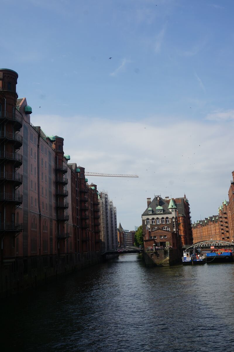 Een foto van een kanaal met aan weerszijden historische bakstenen gebouwen. Aan de linkerkant staan hoge pakhuizen, deels in de schaduw. Aan de rechterkant is een markant gebouw met een groen dak zichtbaar, met daarachter meer bakstenen gebouwen en een brug. Boven het kanaal is een blauwe lucht met enkele wolken en een kraan in de verte te zien.