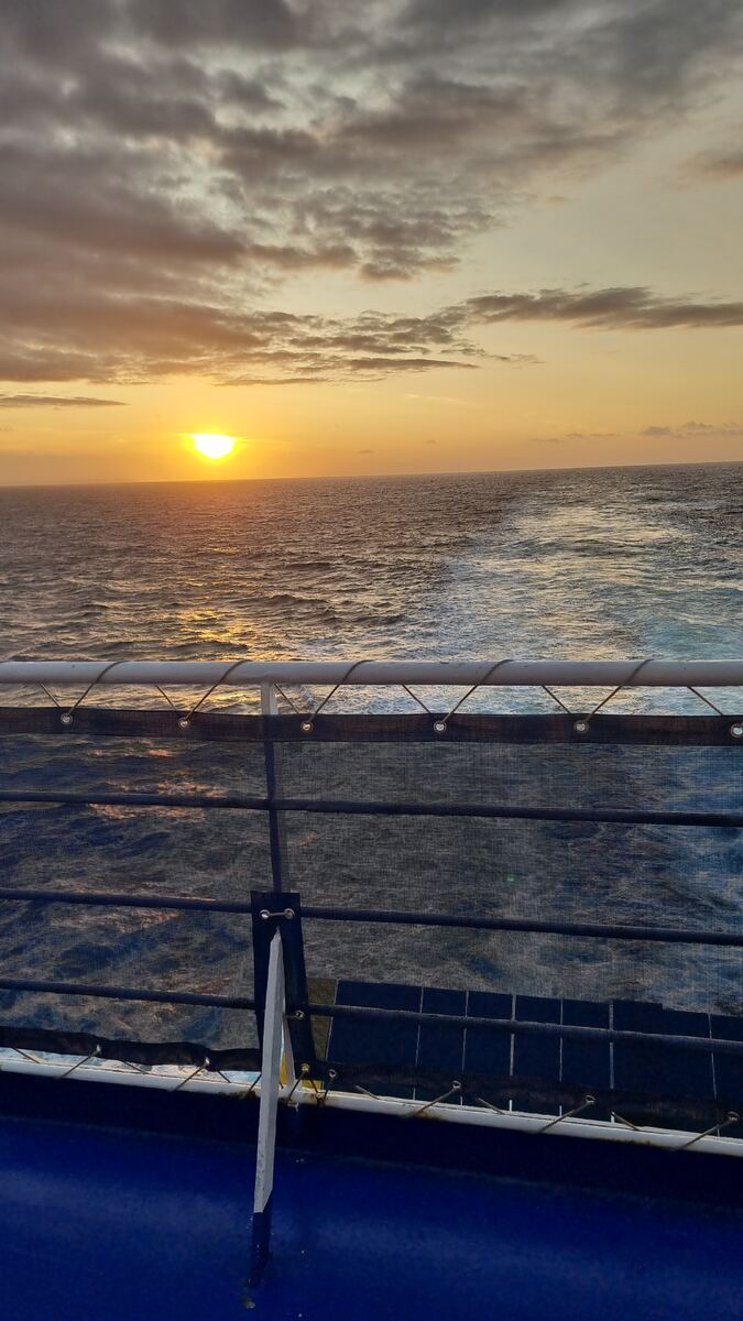 De foto toont een zonsondergang boven een kalme zee, gezien vanaf een schip. De zon is een heldere oranje bol aan de horizon, die het wateroppervlak verlicht met een gouden gloed. De lucht erboven is gevuld met wolken die variëren van donkergrijs tot lichtoranje. Op de voorgrond is een deel van het schip zichtbaar, met een blauwe romp en een reling met een zwart netwerk.