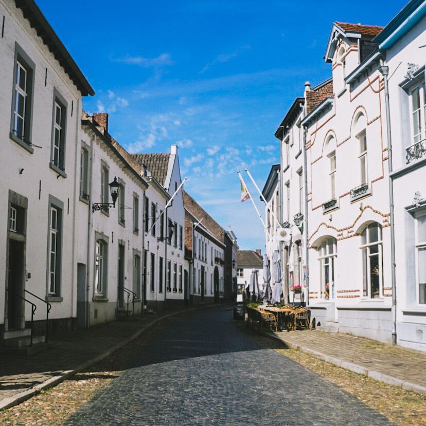 Een smalle straat met kinderkopjes, omzoomd door witgepleisterde gebouwen met traditionele architectuur. Aan de rechterkant is een terras met tafels en stoelen te zien, met parasols die schaduw bieden. De lucht is helderblauw met enkele lichte wolken.