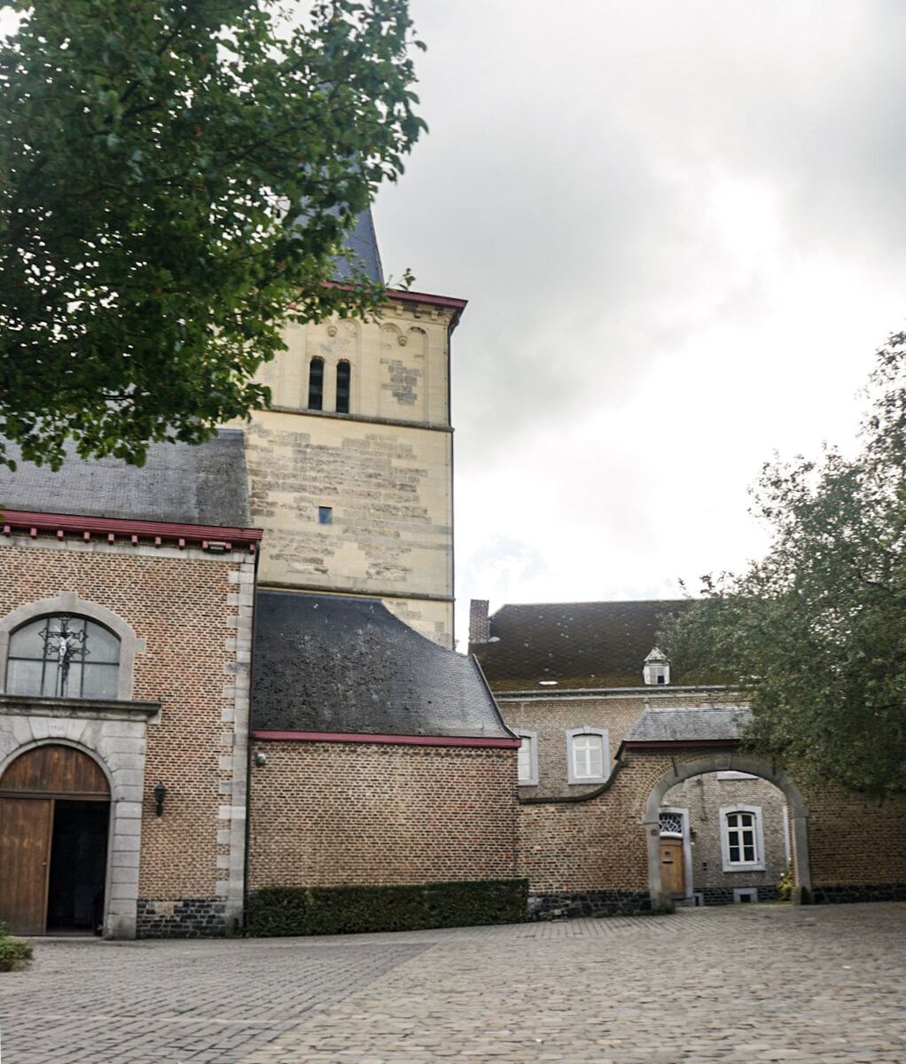 Een foto van een geplaveid plein met aan de linkerkant een bakstenen gebouw met een grote houten deur en een rondboograam. Daarachter staat een hogere, lichtere stenen toren met smalle ramen. Aan de rechterkant is een doorgang onder een stenen boog te zien, die leidt naar een ander gebouw. Boven de gebouwen is een bewolkte lucht zichtbaar en aan de randen van de foto zijn boomtoppen te zien.