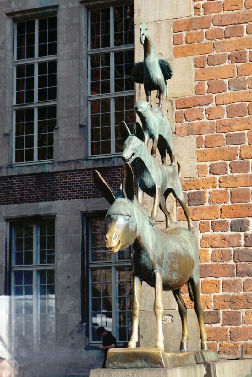 Een bronzen beeld van de Bremer stadsmuzikanten, bestaande uit een ezel, hond, kat en haan, gestapeld op elkaar. Het beeld staat voor een gebouw met bakstenen muren en ramen met roedenverdeling.