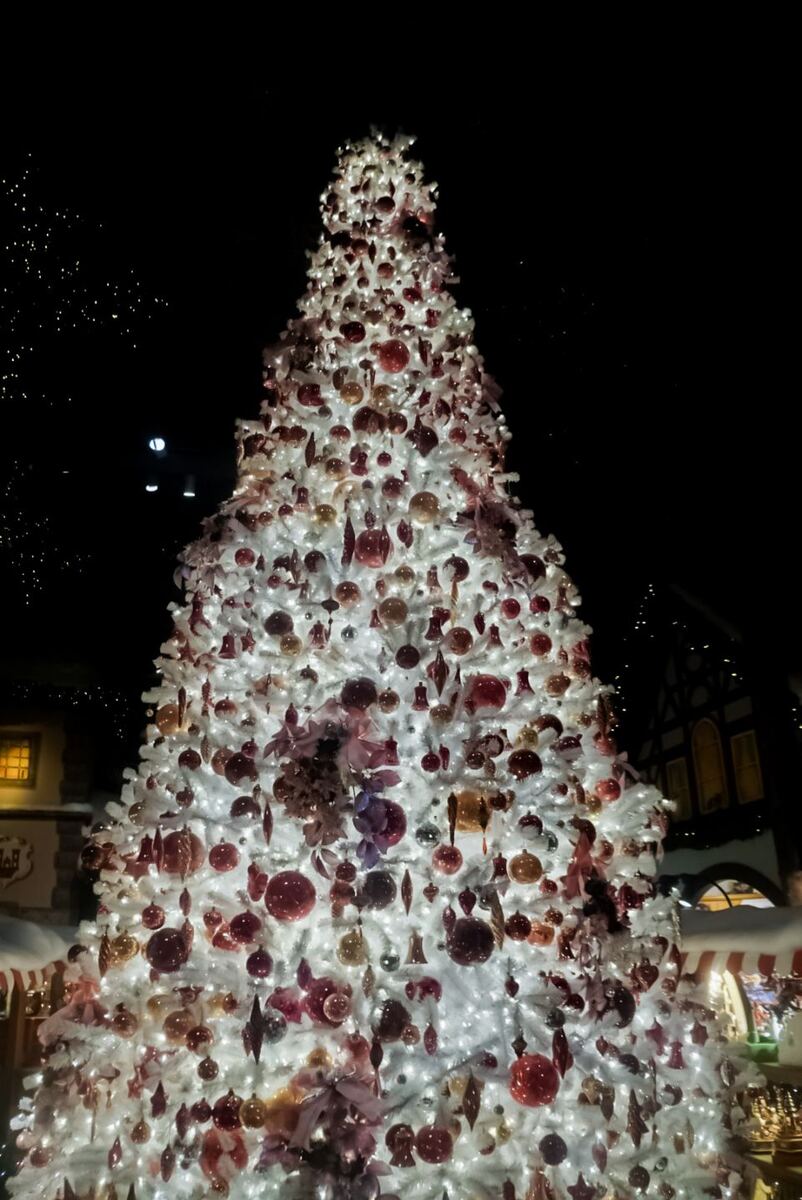 Een grote, wit verlichte kerstboom met rode en gouden kerstballen en decoraties, staand tegen een donkere achtergrond. Aan de zijkanten zijn delen van gebouwen met verlichte ramen zichtbaar.