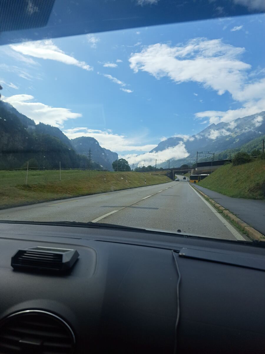 De foto is genomen vanuit de voorruit van een auto, met een deel van het dashboard zichtbaar aan de onderkant. Een tweebaansweg met witte belijning strekt zich uit naar de horizon, omzoomd door groene grasbermen. Aan de rechterkant van de weg is een gele bus te zien, en verderop rijden enkele andere voertuigen. De weg loopt onder een viaduct door, waarboven spoorlijnen met bovenleidingen zichtbaar zijn. Links en rechts van de weg verheffen zich imposante, met bomen begroeide bergen, waarvan somm