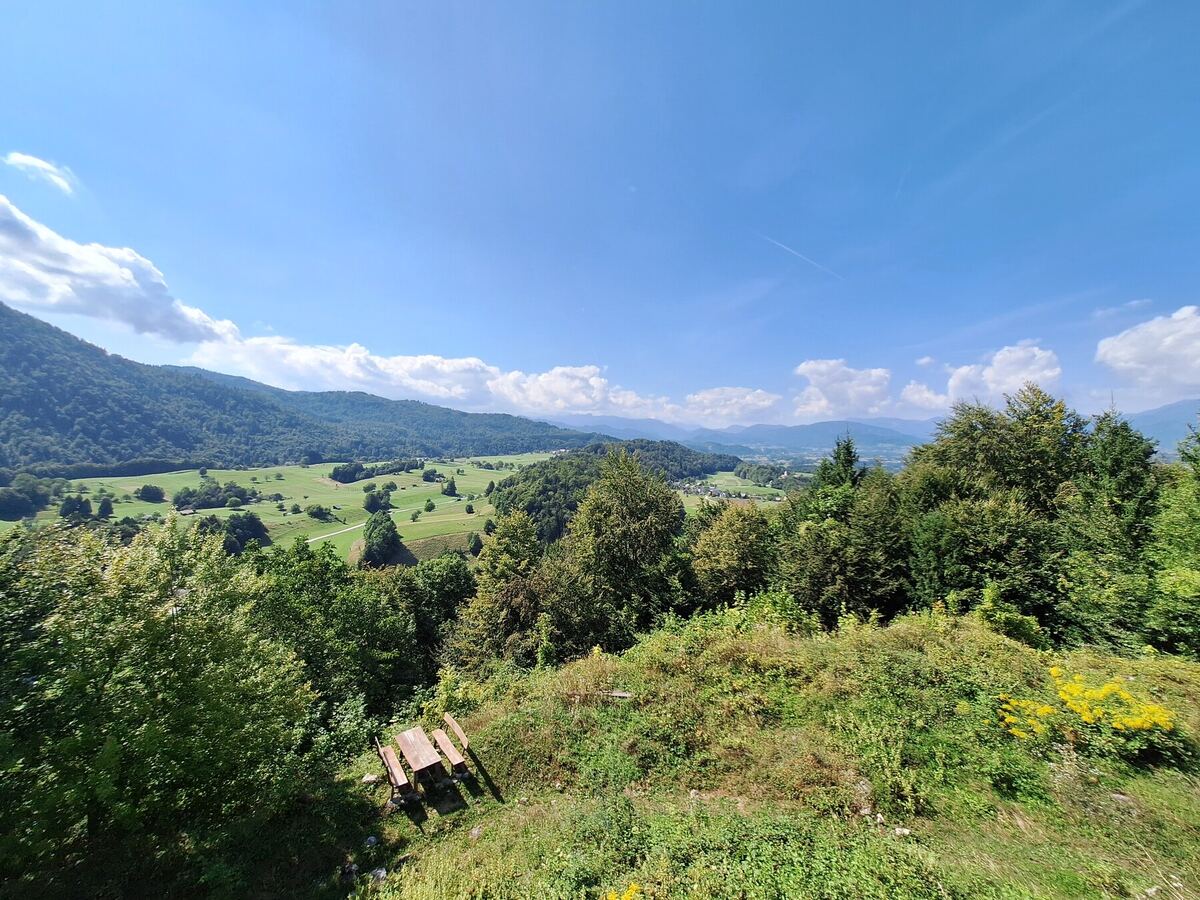 Een weids uitzicht over een groene vallei met glooiende heuvels, bossen en verspreide huizen onder een helderblauwe lucht met enkele witte wolken. Op de voorgrond is een houten picknicktafel met banken zichtbaar, omringd door dichte begroeiing.