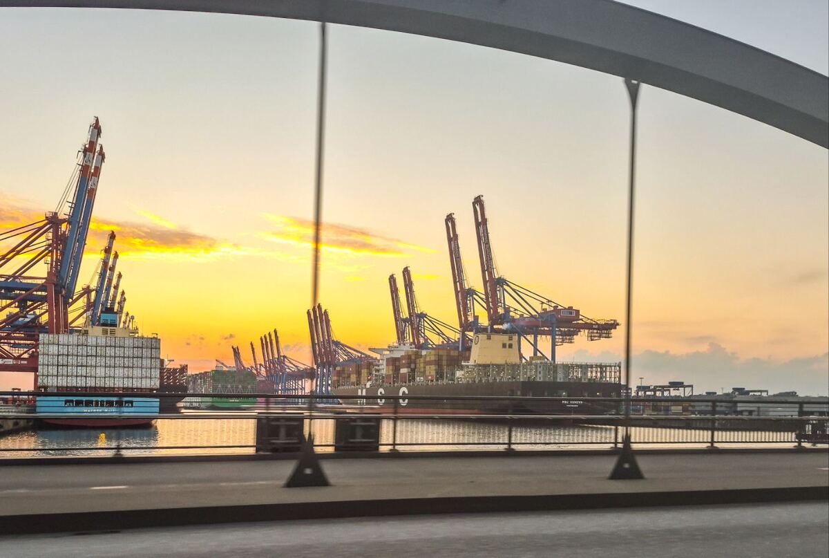 Een havenlandschap bij zonsondergang, gezien door de constructie van een brug. Grote containerkranen en vrachtschepen liggen aan het water, met de oranje gloed van de ondergaande zon aan de horizon.