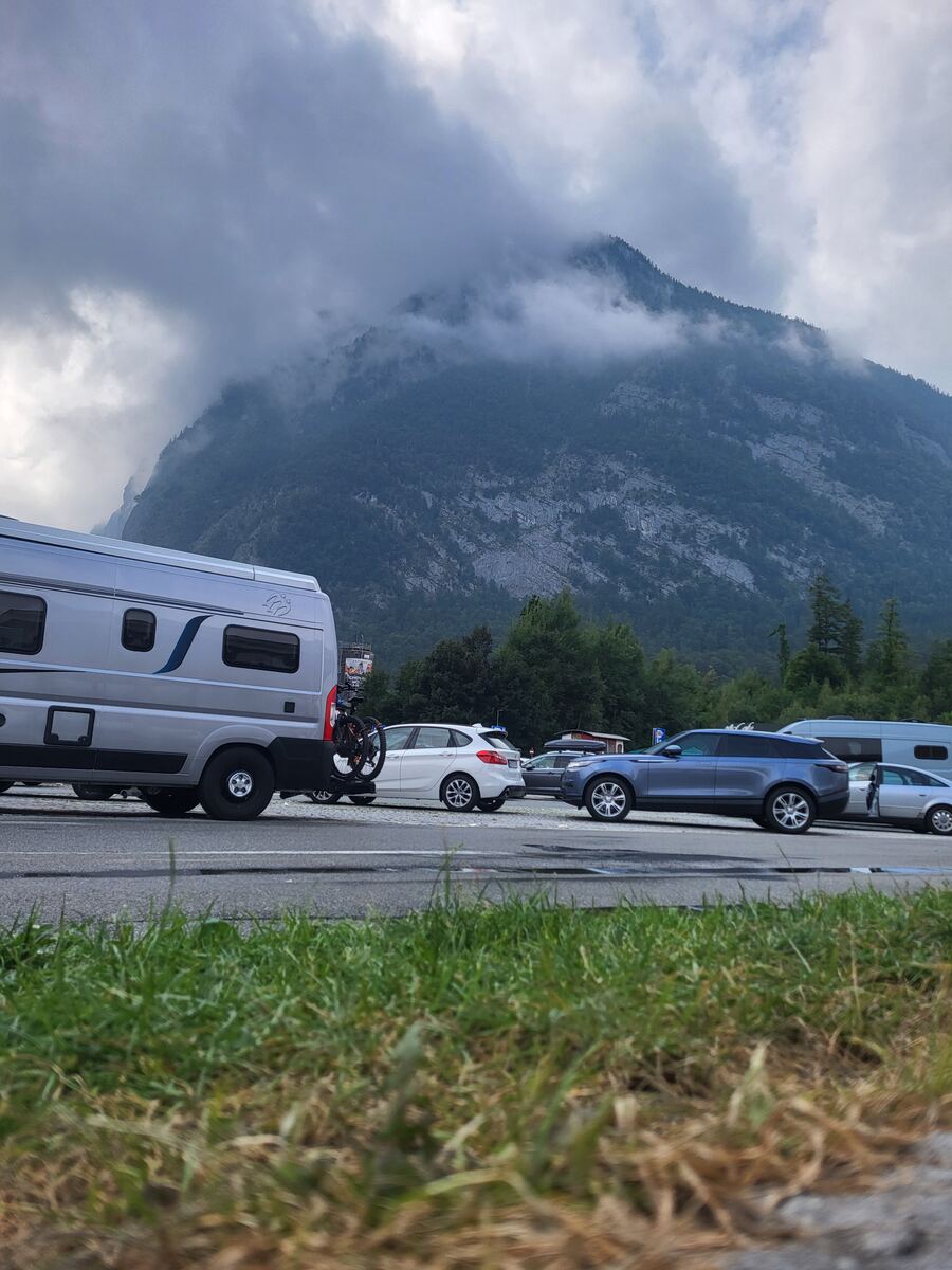 Een parkeerplaats met verschillende voertuigen, waaronder een camper met fietsenrek en een fiets, met op de achtergrond een grote berg bedekt met wolken en groen. De voorgrond toont gras en een deel van de weg.