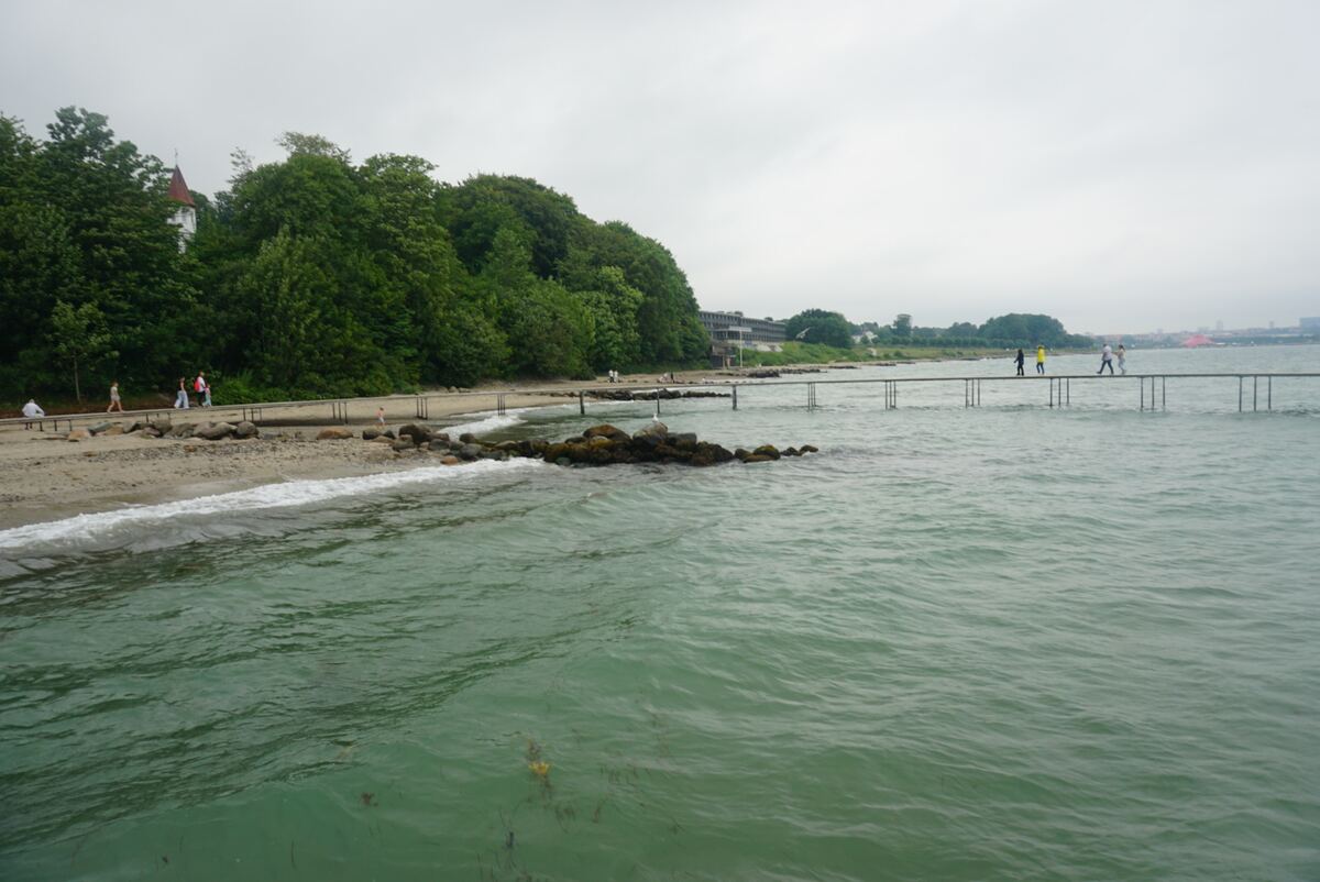 Een kustlandschap met een zandstrand, rotsen in het water, en een houten promenade langs de kust. Op de achtergrond is een beboste heuvel te zien met een gebouw met een rood dak en een groter gebouw. Verderop in het water loopt een lange, smalle steiger met mensen erop. De lucht is bewolkt en het water is groenachtig van kleur.