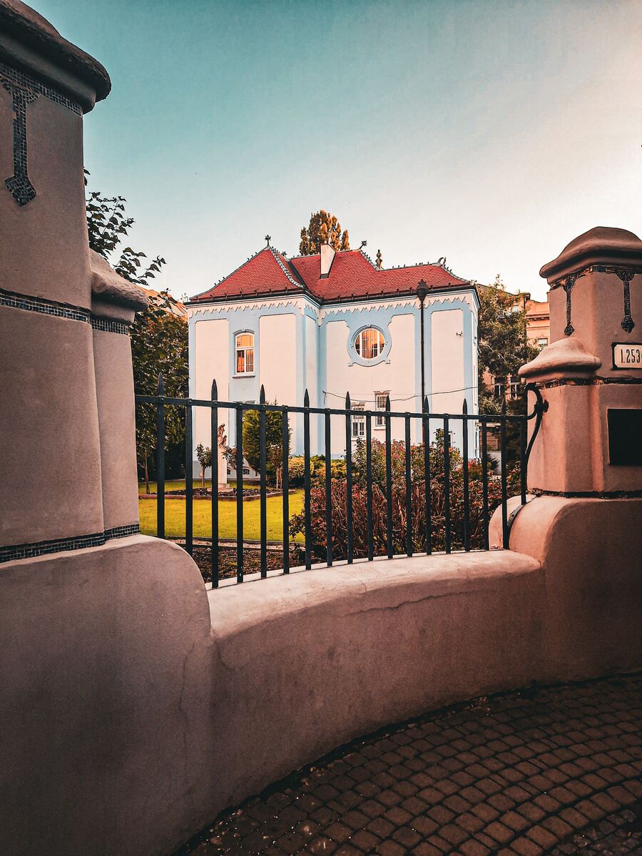 Een wit huis met een rood dak en lichtblauwe accenten, gezien door een zwart hek. De gevel heeft een rond raam en er is een groene tuin voor het huis. Op de voorgrond zijn delen van een stenen muur met decoratieve tegels zichtbaar. De lucht is lichtblauw met een oranje gloed aan de horizon.