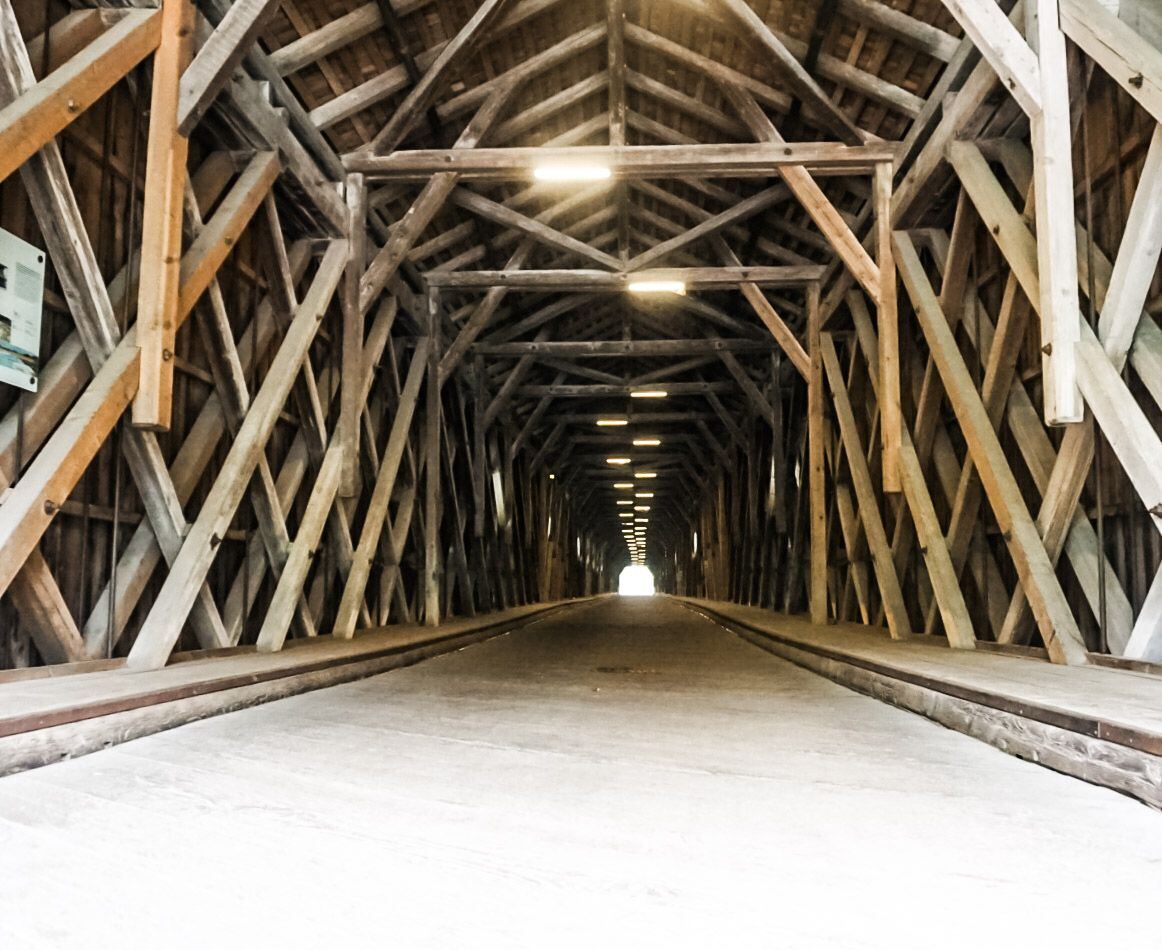Een lange, overdekte houten brug met een constructie van kruisende balken aan de zijkanten en het plafond. In het midden van de brug is een rij lampen zichtbaar die de weg verlichten. Aan het einde van de brug is een heldere opening te zien, wat het einde van de tunnel aangeeft. De vloer van de brug is van beton of asfalt.