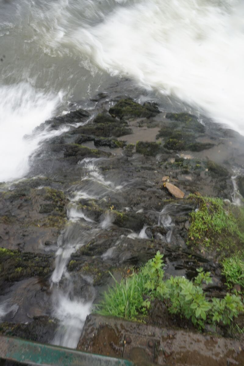 Een close-up van snelstromend water dat over donkere, met mos begroeide rotsen en stenen stroomt. Het water is deels wazig door beweging, wat de snelheid benadrukt. Rechtsonder is een stukje groen gras en een deel van een houten of metalen constructie zichtbaar.