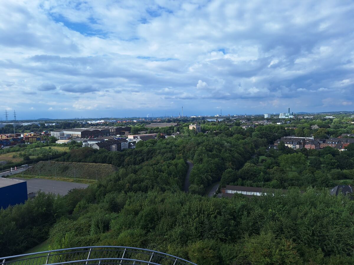 Een weids uitzicht over een stedelijk landschap met veel groen, industriële gebouwen en woonhuizen onder een bewolkte hemel. Op de voorgrond is een deel van een metalen reling zichtbaar.