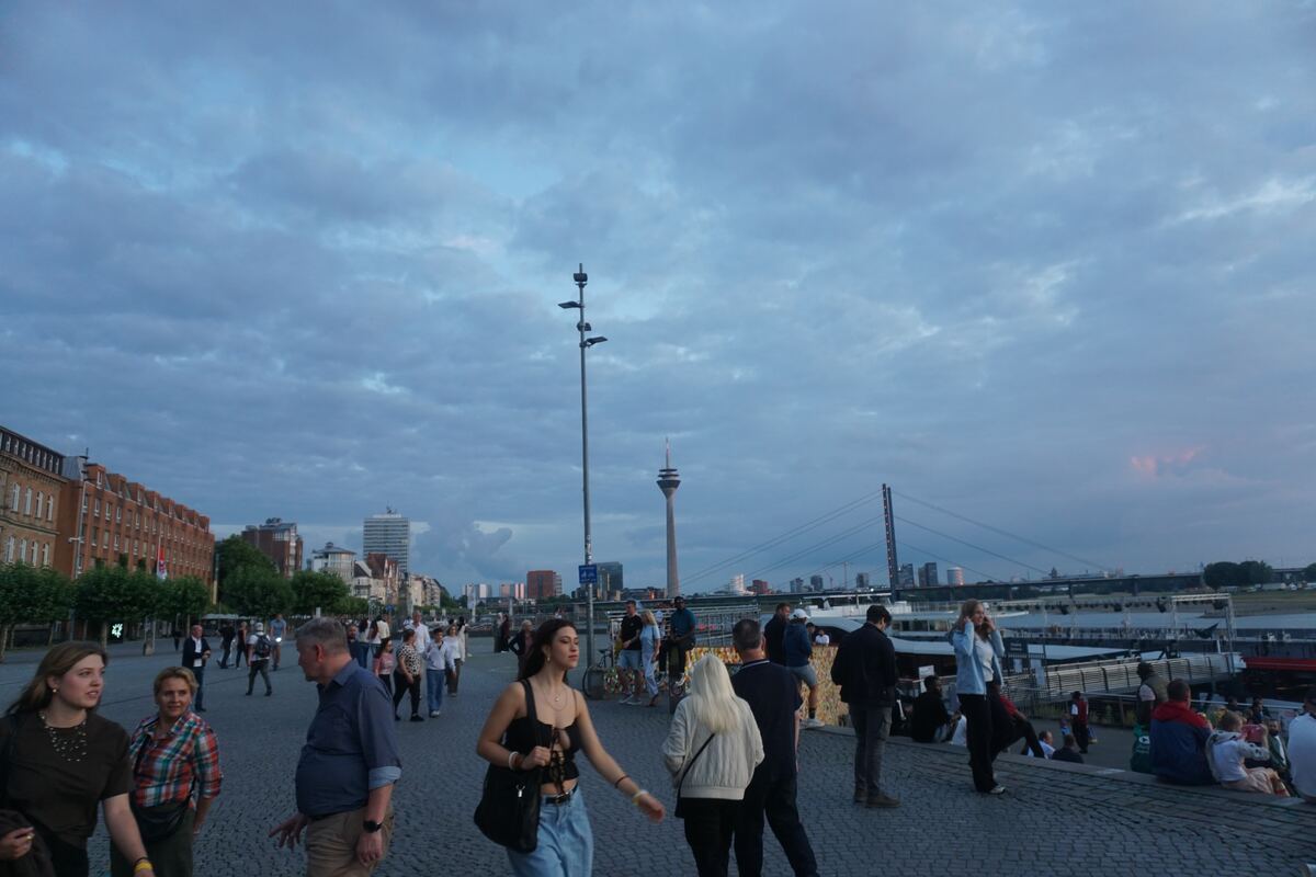 Een avondbeeld van een kade langs een rivier, met mensen die wandelen en zitten. Op de achtergrond zijn gebouwen, een brug en een hoge zendmast zichtbaar onder een bewolkte lucht.