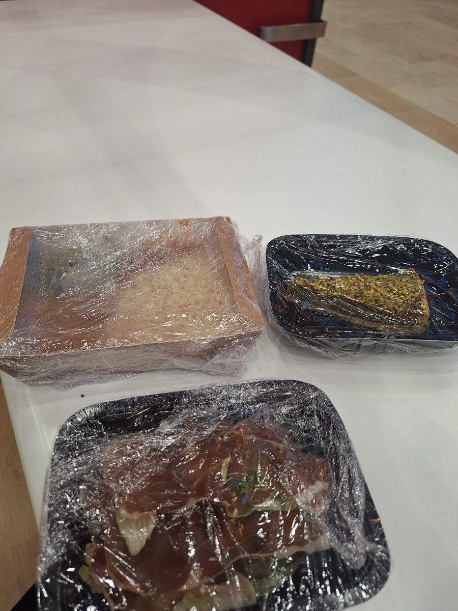 Drie voorverpakte maaltijden, verpakt in plasticfolie, liggen op een witte tafel. Eén maaltijd zit in een houten bakje en lijkt rijst te bevatten. De andere twee liggen in zwarte plastic bakjes, waarvan één met een gerecht bedekt met groene korrels en de andere met vleeswaren.