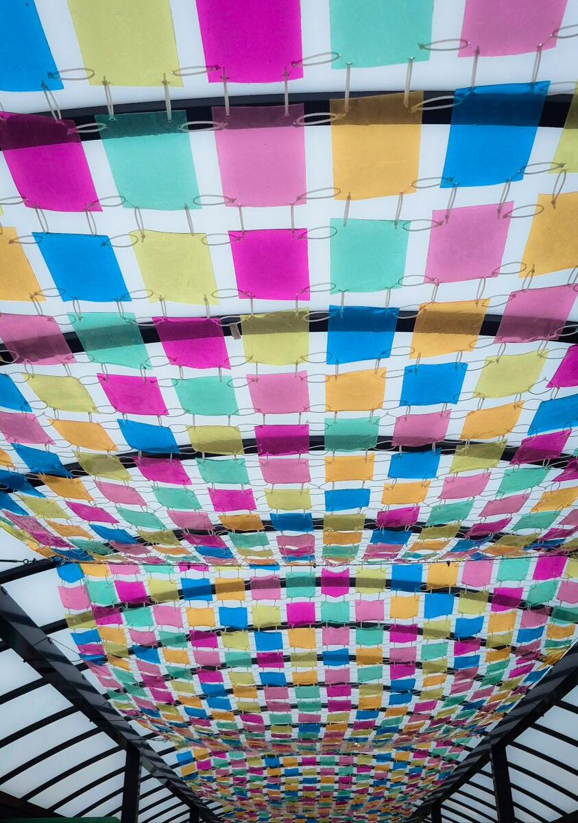 Een plafond bedekt met een raster van kleine, gekleurde vierkante lapjes stof of plastic in de kleuren roze, geel, blauw en groen. De lapjes zijn bevestigd aan een netwerk van draden en hangen onder een dakconstructie met donkere balken.