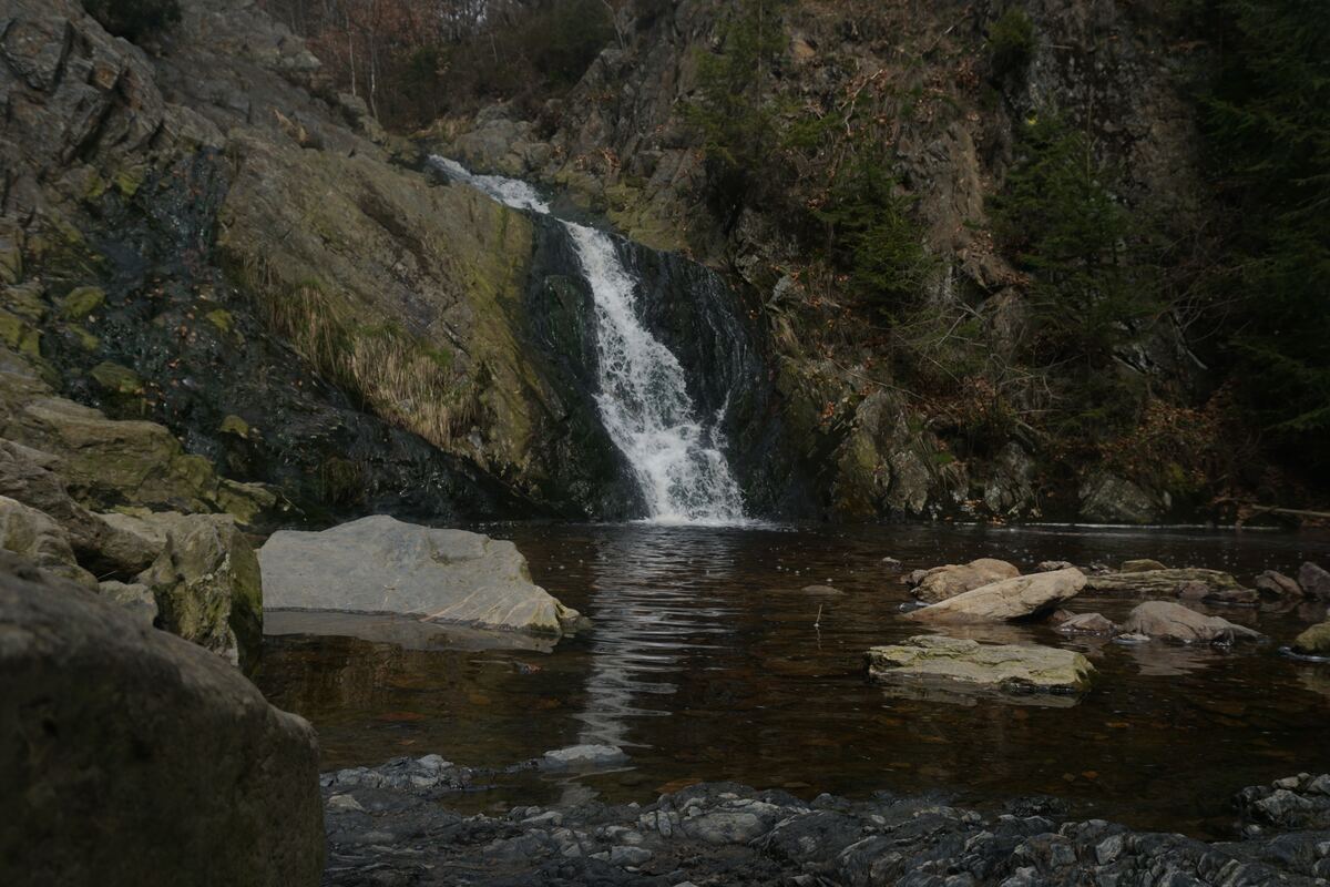Een waterval stroomt over een rotswand in een poel met donker water. Rondom de waterval zijn rotsen en wat begroeiing te zien. De lucht is bewolkt en de algehele indruk is van een natuurlijke, rustige omgeving.