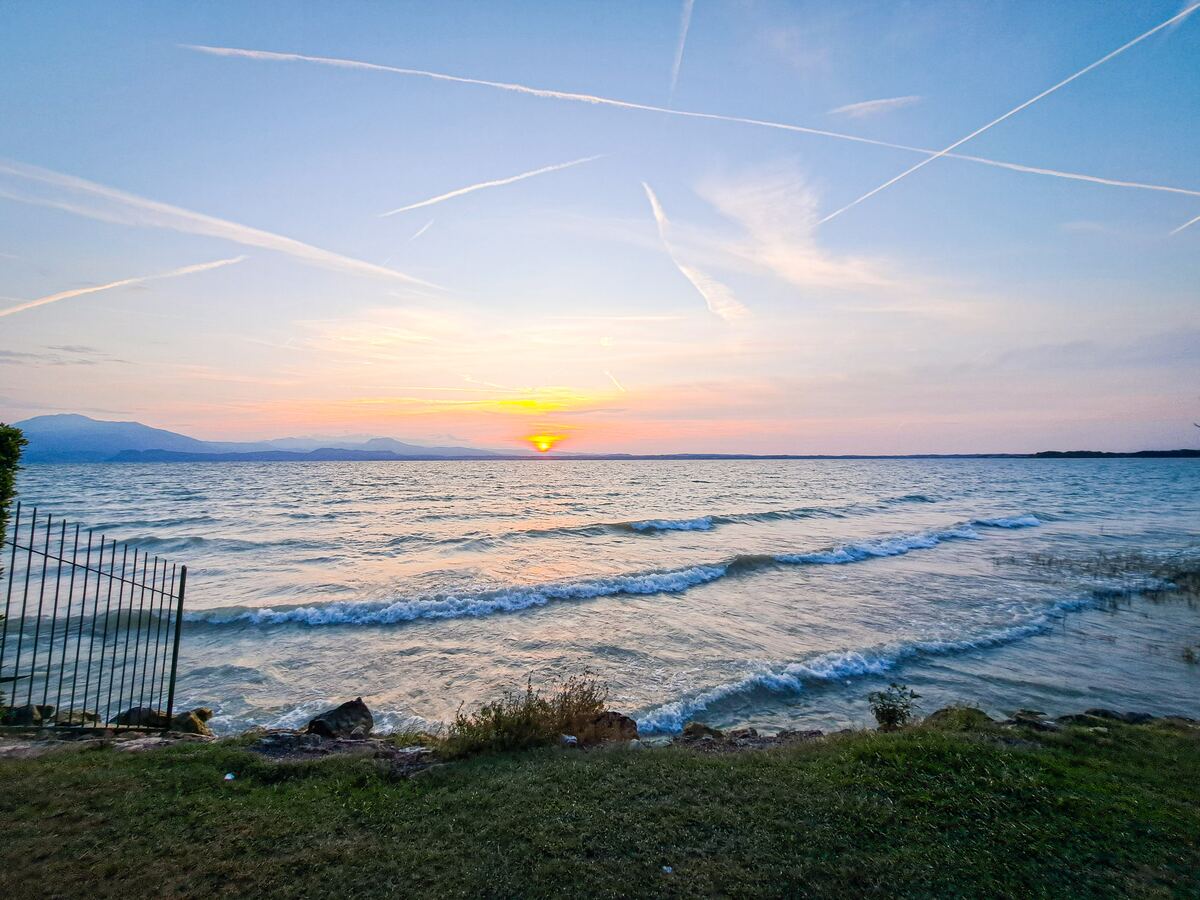 Een zonsopkomst boven een groot meer met golven die de oever bereiken. Aan de horizon zijn bergen zichtbaar en de lucht is gevuld met vliegtuigstrepen. Op de voorgrond is een stukje gras en een metalen hek te zien.