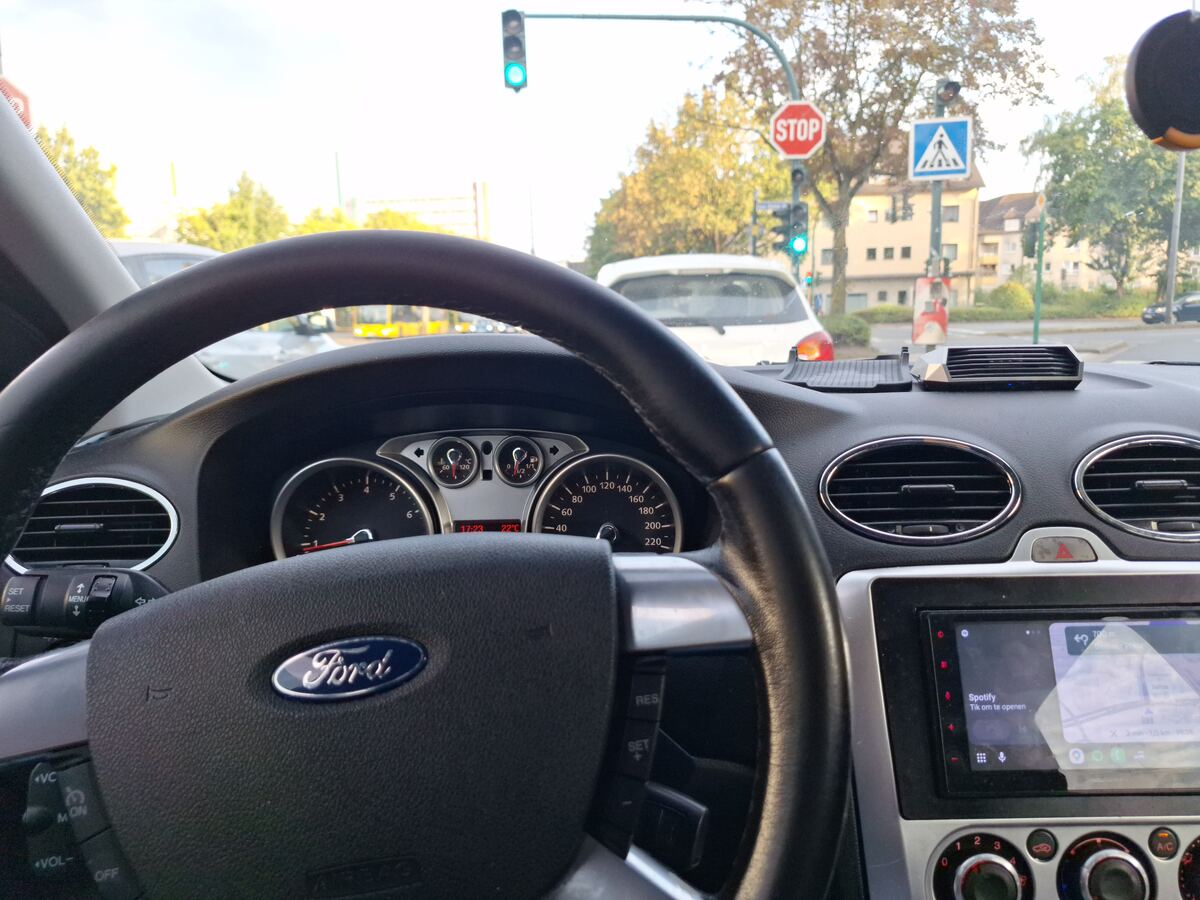 Een interieurfoto van een Ford Focus met het stuur en dashboard op de voorgrond. Door de voorruit is een stadsbeeld te zien met een verkeerslicht op groen, een stopbord en een zebrapadbord. Er rijden andere auto's en gebouwen zijn zichtbaar op de achtergrond. Op het dashboard is een navigatiesysteem met Spotify te zien.
