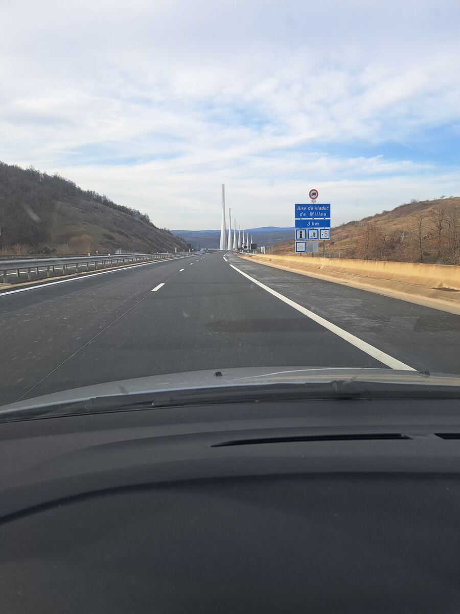 Een foto genomen vanuit een auto op een snelweg, met op de achtergrond de kenmerkende pilaren van het Viaduct van Millau. Rechts van de weg staat een blauw verkeersbord met de tekst 'Aire du viaduc de Millau 3 km' en symbolen voor informatie, een picknickplaats en een uitzichtpunt. De weg is omgeven door heuvelachtig, deels bebost landschap onder een bewolkte lucht.