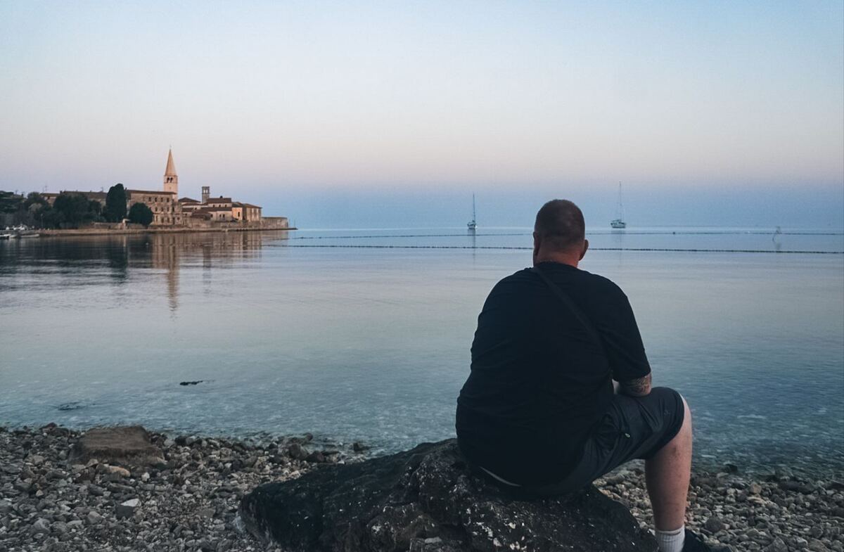Een persoon zit op een rotsblok aan een kiezelstrand en kijkt uit over het water. Aan de overkant van het water is een historische stad te zien met een opvallende kerktoren. Op het water liggen enkele zeilboten en de lucht is lichtblauw met een zachte roze tint.