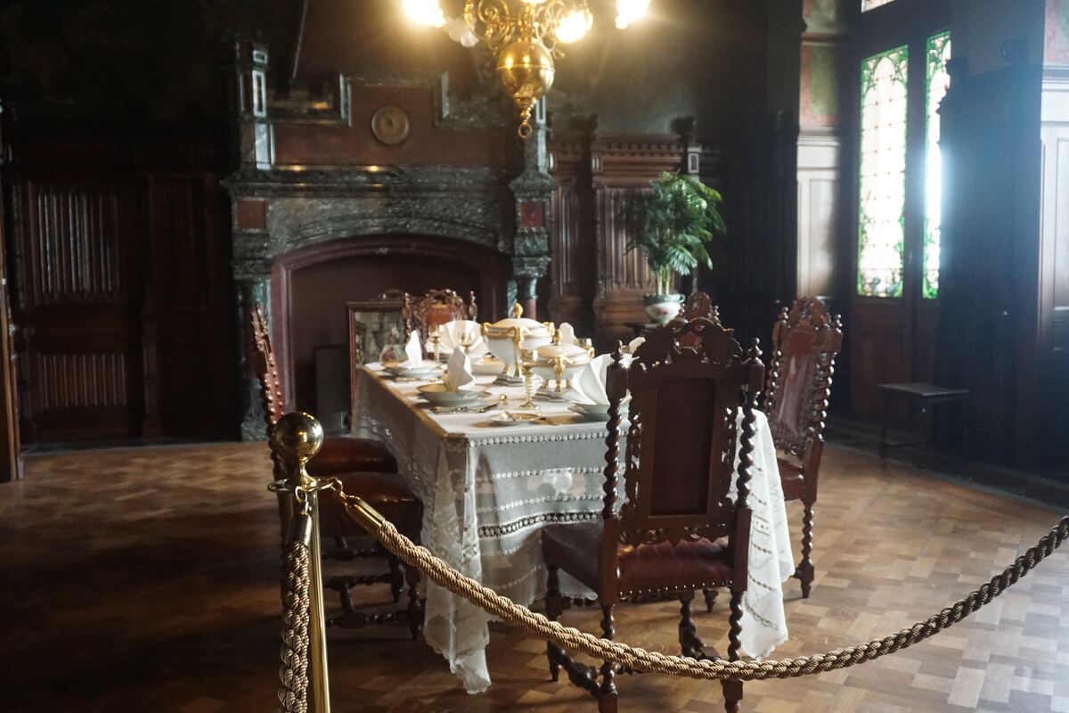 Een rijk gedekte eettafel met wit linnen, servies en bestek, omringd door donkerhouten stoelen in een historische eetkamer. Op de achtergrond is een grote open haard te zien en een raam met glas-in-lood. Een gouden kroonluchter hangt boven de tafel en een afzetkoord scheidt de tafel van de kijker.